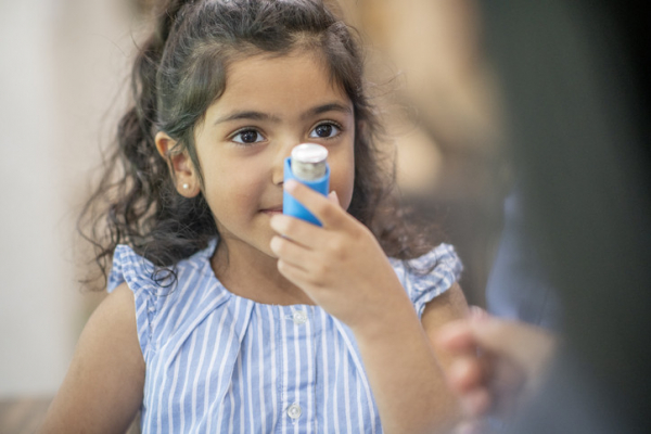 Child with dark hair and eyes wearing a blue and white striped top is learning how to use an asthma inhaler, which she holds near her mouth; blurry adult seen partially from the back